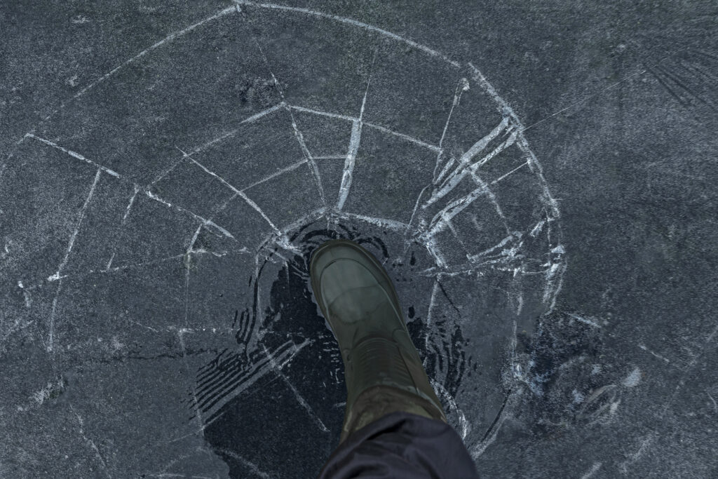 Fisherman foot on broken cracked thin ice at lake. Dangerous fishing